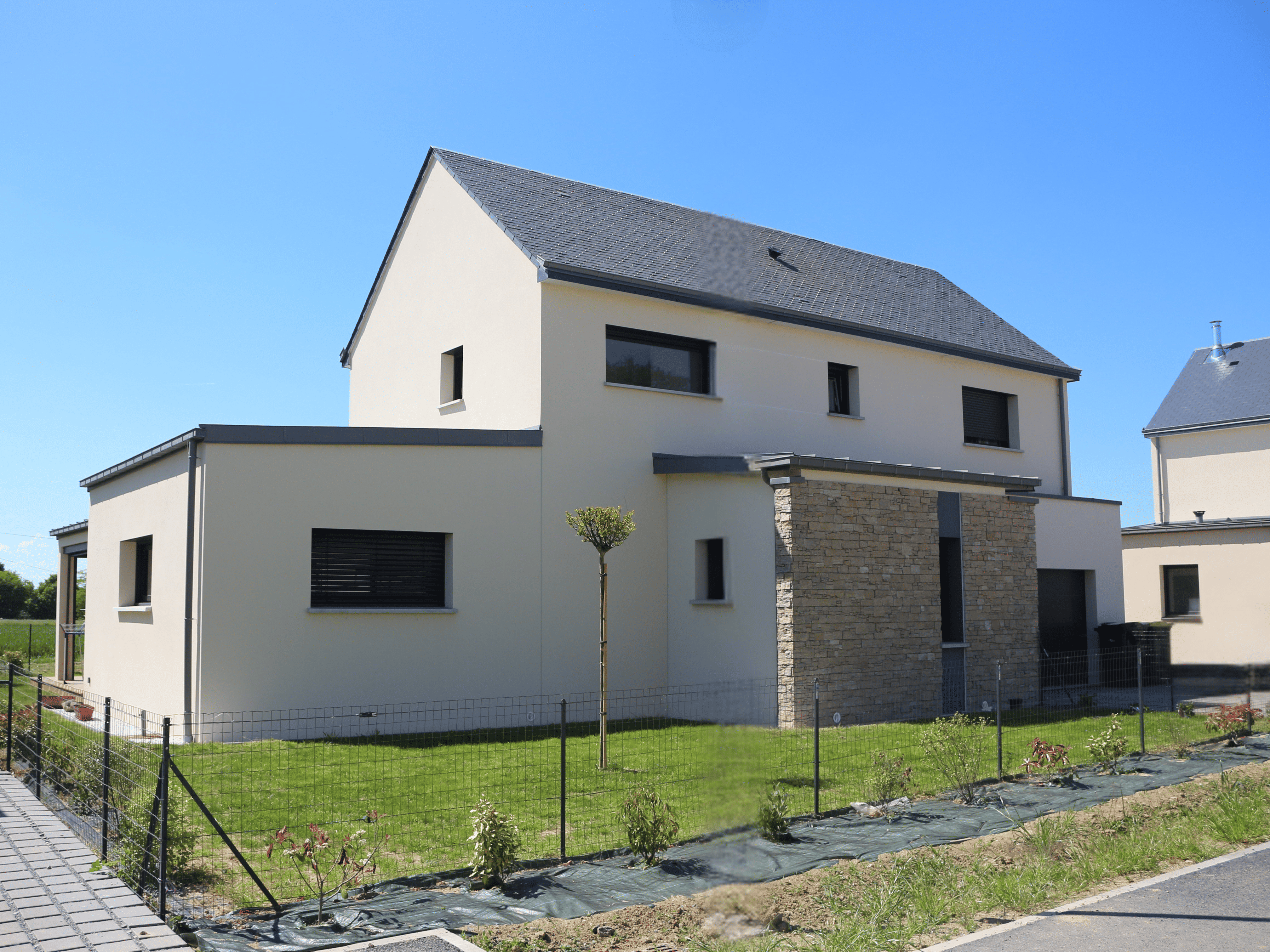 Maison contemporaine à étage avec toit en double pente, façade en enduit clair et parement pierre, menuiseries noires et jardin paysager Maison contemporaine à étage avec toit en double pente, façade en enduit clair et parement pierre, menuiseries noires et jardin paysager
